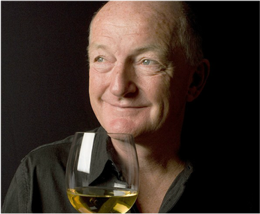 Close-up of a man holding a glass of white wine, smiling, with a dark background.