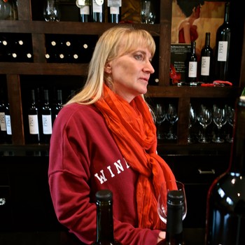 Woman in red sweater and orange scarf at Lodi Winegrape Commission event with wine bottles in background.