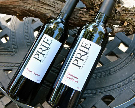Two bottles of Prie wine from Lodi Winegrape Commission, Petit Verdot and Cabernet Sauvignon, on a black metal surface.
