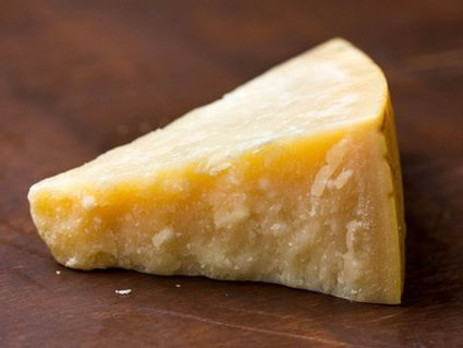 Close-up of a wedge of hard cheese on a wooden table, showing its textured surface and pale yellow color.