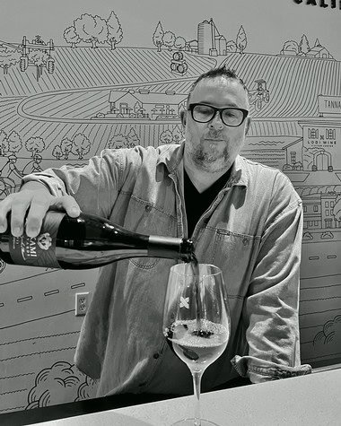 Man pouring wine into a glass at Lodi Winegrape Commission event, with a mural background of vineyards and wineries.