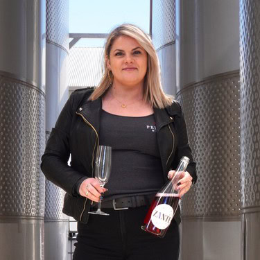 Woman holding a wine glass and bottle at a winery with stainless steel tanks in the background.