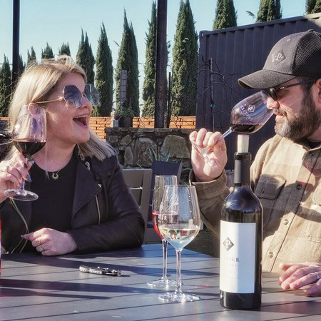 Two people enjoying wine tasting outdoors with a bottle of wine and glasses, surrounded by cypress trees.