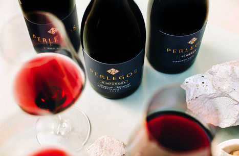 Three bottles of Perlegos wine and two glasses of red wine on a table with rocks.
