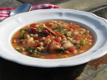 A bowl of hearty bean and vegetable soup with tomatoes, greens, and herbs, served on a rustic outdoor table.