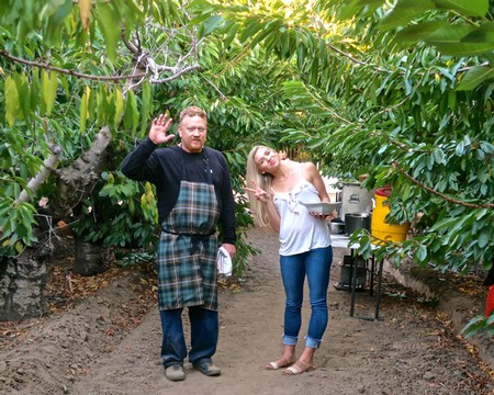Two people in a vineyard, one waving, the other holding a tray, surrounded by lush grapevines.