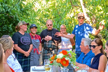 Group of people at outdoor event with wine and flowers, under lush green trees, discussing wine grapes.