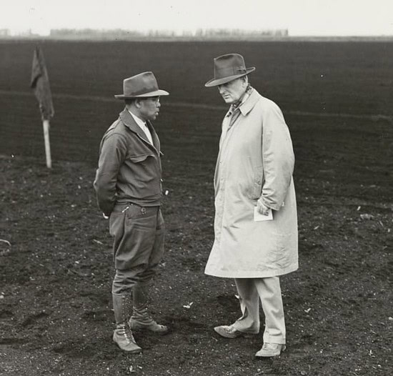 Two men in vintage clothing and hats examining vineyard soil in a field.