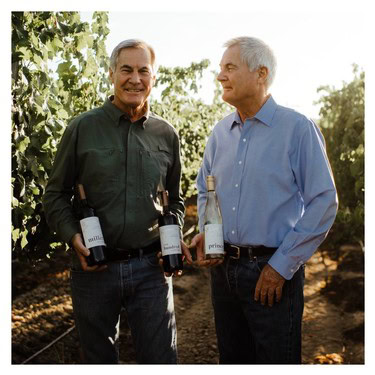 Two men in vineyard holding wine bottles, smiling, during daytime in Lodi wine region.