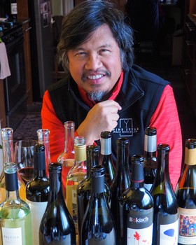 Man smiling at wine tasting event with various wine bottles in front of him.