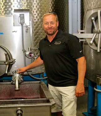 Man in black polo shirt standing in wine production facility with stainless steel tanks.