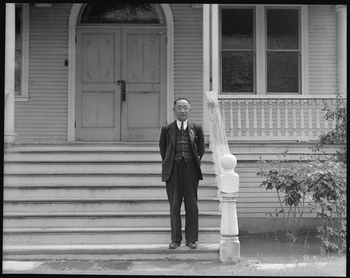 Man in formal attire standing on front steps of a vintage house with wooden stairs and railing.