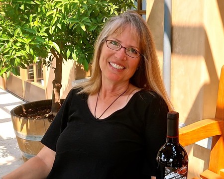 Smiling woman with glasses sitting outdoors with a bottle of wine, greenery, and wooden furniture in the background.