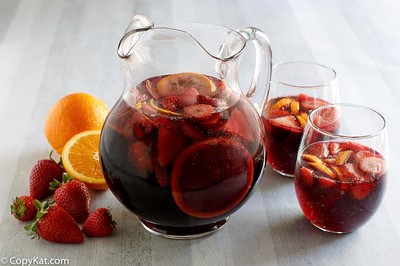 Glass pitcher of red wine with strawberries, oranges, and ice, alongside fresh strawberries and oranges on a light surface.