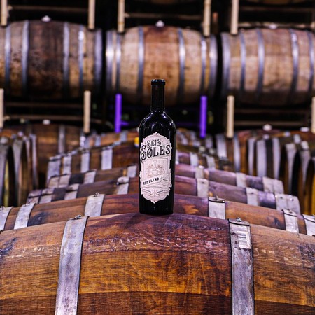 Wine bottle labeled 'Seis Soles' on wooden barrels in a winery cellar.