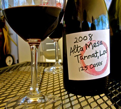 Glass of red wine with a bottle labeled 2008 Alta Mesa Tannat Lodi, California, on a metal table.