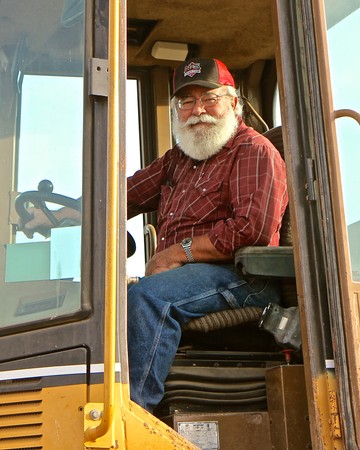Smiling man with a beard and glasses in a red plaid shirt and cap sitting in a tractor cab.