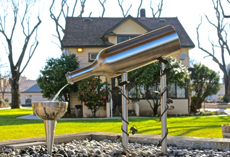 Artistic wine bottle fountain in Lodi with flowing water, surrounded by greenery and a house in the background.