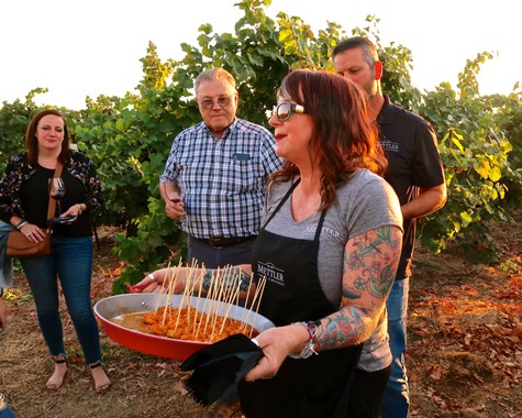 Group of people at a vineyard tasting event, woman holding a tray of wine samples, lush grapevines in the background.