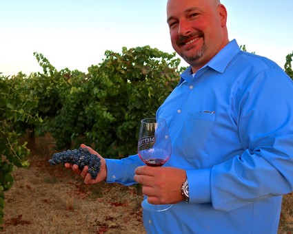 Man holding a bunch of grapes and a glass of wine in a vineyard during sunset.