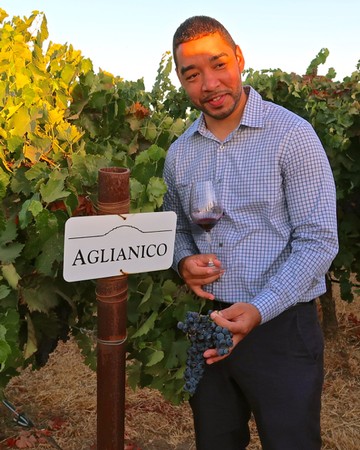 Man tasting wine in a vineyard with a glass of red wine and a bunch of grapes, near a sign that says 'Aglianico'.