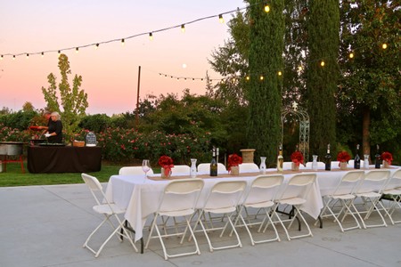 Elegant outdoor wine tasting setup with white tablecloths, wine bottles, and string lights at sunset in Lodi, California.