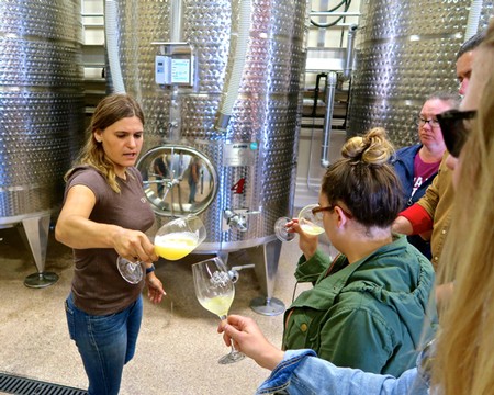 Group tasting wine at a winery with large stainless steel fermentation tanks in the background.