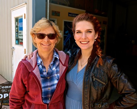 Two women smiling outdoors at a Lodi Winegrape Commission event, one wearing sunglasses and a red jacket, the other in a blue.
