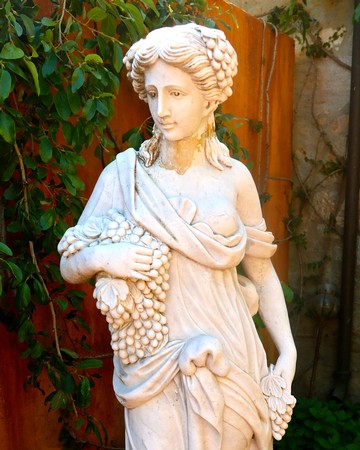 Marble sculpture of a woman holding grapes, symbolizing wine and harvest, set outdoors with greenery and wooden fence.