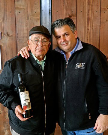 Two men at a wine event, one holding a wine bottle, in front of a wooden background.