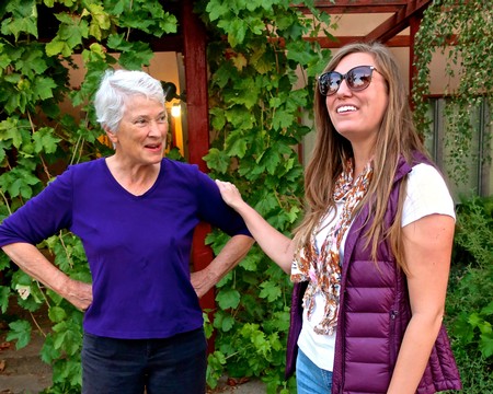 Two women talking outdoors among grapevines, one older and one younger, smiling and engaging in conversation.