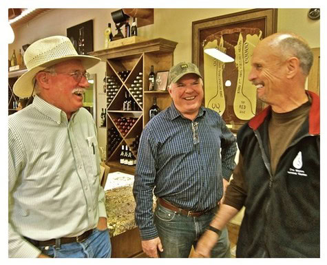 Three men smiling and talking inside a wine shop, with wine bottles and artwork in the background.