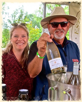 Smiling woman and man at a wine tasting event, man holding a wine bottle, outdoor setting with trees.