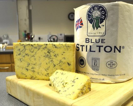 Blue Stilton cheese with distinctive blue veins on a wooden cutting board, with packaging in the background.