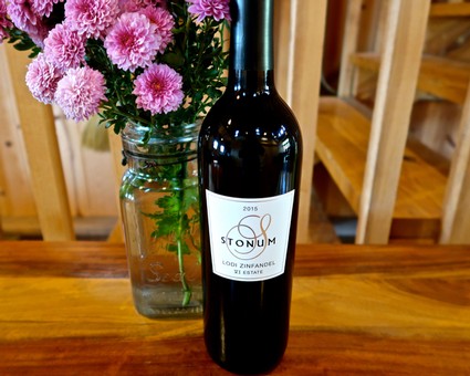 A bottle of Lodi Zinfandel wine from Stonum, placed on a wooden table with a vase of pink flowers in the background.
