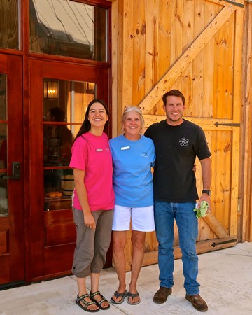 Three smiling people standing outside a wooden building at a Lodi Winegrape Commission event.