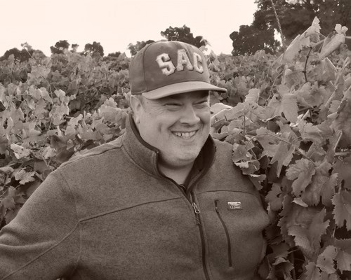 Smiling man in vineyard wearing a cap and jacket, surrounded by grapevines, promoting Lodi Winegrape Commission.