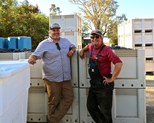 Two workers in vineyard gear smiling next to grape bins at Lodi Winegrape Commission.