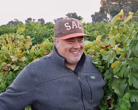 Man smiling in vineyard with lush grapevines during daytime in Lodi, California.