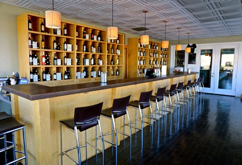 Modern tasting room with wine bottles on wooden shelves and a long bar with chairs for wine tasting.