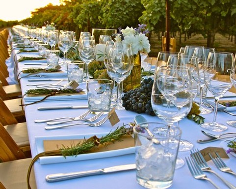 Elegant outdoor dining table set in a vineyard with wine glasses, floral centerpieces, and grape bunches for a wine event.