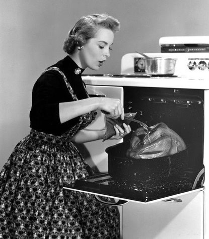 Black and white photo of a woman placing food in an oven, vintage kitchen scene from mid-20th century.