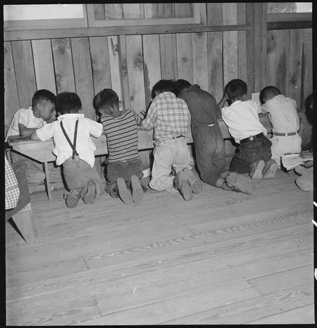 Children kneeling and praying in a line inside a wooden room, engaged in a religious activity.