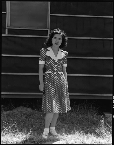 Black and white photo of a woman in a polka dot dress standing outdoors near a wooden structure.