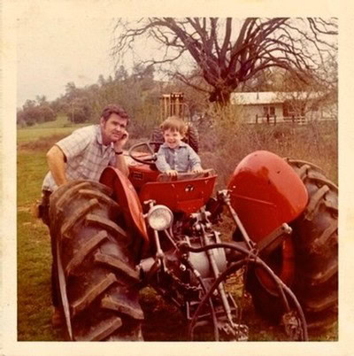 Man and child on a vintage red tractor in a rural setting with a large tree in the background.
