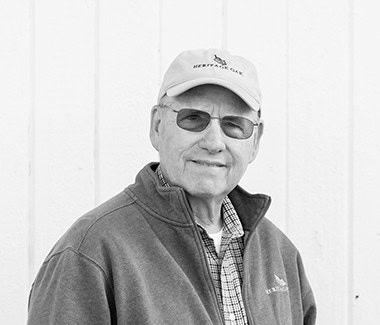 Older man wearing sunglasses and a baseball cap, smiling outdoors in front of a white wall.