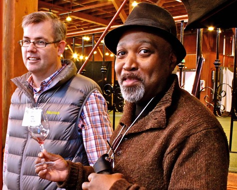 Two men at a wine tasting event, one holding a wine glass, in a rustic setting with warm lighting.