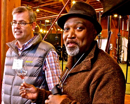 Two men at a wine event, one holding a wine glass, in a rustic setting with warm lighting.