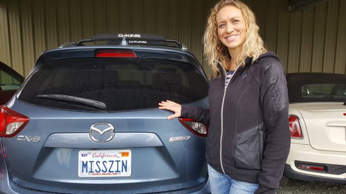 Smiling woman standing next to a Mazda SUV with a personalized license plate reading 'MISSZYN'.