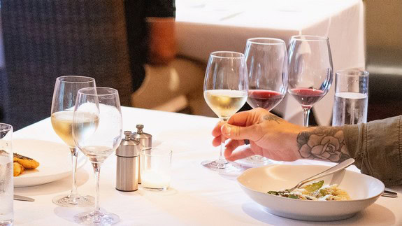 Person holding a glass of white wine during a wine tasting event with multiple wine glasses on a table.